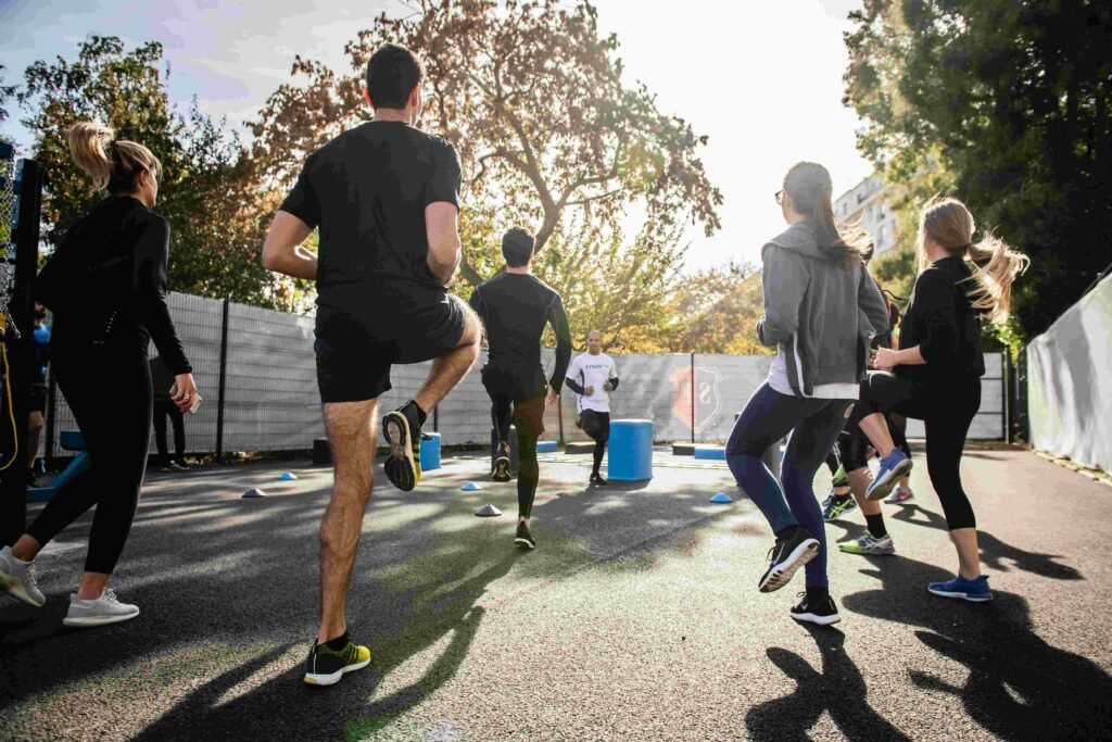 A group of people participating in an outdoor fitness workout session in a park during sunny morning hours, performing high-knee exercises under the guidance of a trainer.